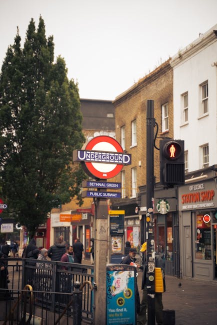 A street scene in Bethnal Green featuring a brick building with large arched windows on the left side, and an old railway bridge with a yellow and green 'Camden Lock' sign extending across the road. I