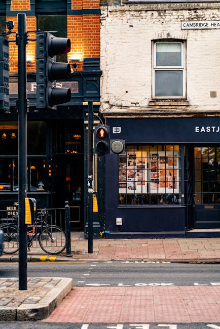 A view of a busy street scene outside a building on Cambridge Heath Road, Bethnal Green, featuring a traffic light with red signals at the corner of the pavement and a bicycle locked to a black metal bike rack in the foreground. The building has a dark blue storefront with large windows displaying interior lighting and various items, possibly from a cafe or shop. Above the storefront, there is a white brick wall with a window and a street sign reading 'Cambridge Heath Road.' The street is paved with red and grey bricks, and a section of road with white markings is visible near the curb. This scene captures the initial setup phase of a home relocation or furniture transport, with the building likely serving as a point of access for a removals company such as Man with Van Bethnal Green, who coordinate packing and loading processes for house moves.