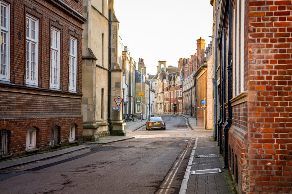 Photograph of a quiet residential street in Bethnal Green, showing a narrow tarmac road flanked by red brick and stone buildings with multiple windows and decorative architectural details. The street features a slight curve with a parked grey car visible near the corner, while bicycles are secured to a railing on the sidewalk. The scene is illuminated by natural daylight, casting soft shadows, and the surroundings include a few street signs and pavement markings. This setting depicts a typical London neighbourhood, relevant for home relocation and furniture transport, as seen in visual material used by Man with Van Bethnal Green for house removals and moving services.