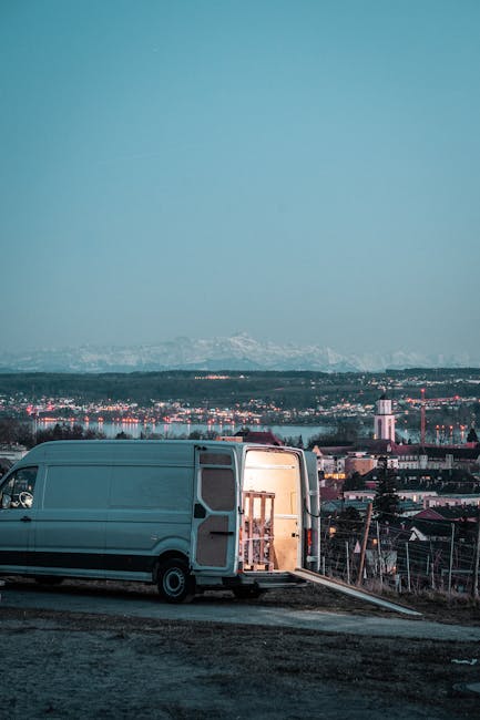 A light grey moving van with its rear doors open, parked on a paved area during twilight hours. Inside the van, a wooden chair and various packing materials are visible, indicating ongoing packing or