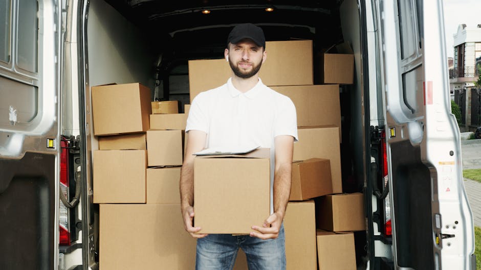 A light grey moving van with its rear doors open, parked on a paved area during twilight hours. Inside the van, a wooden chair and various packing materials are visible, indicating ongoing packing or