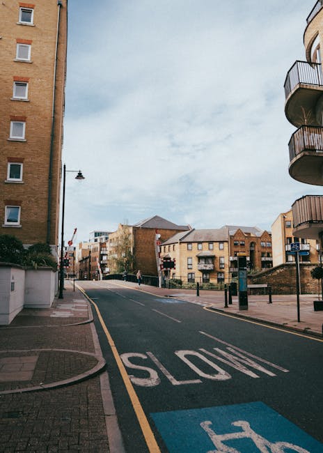 An urban street scene outside a residential building in Bethnal Green with a gently sloping road marked with a 'SLOW' sign in white paint and a designated parking area in blue. The scene shows a mix o